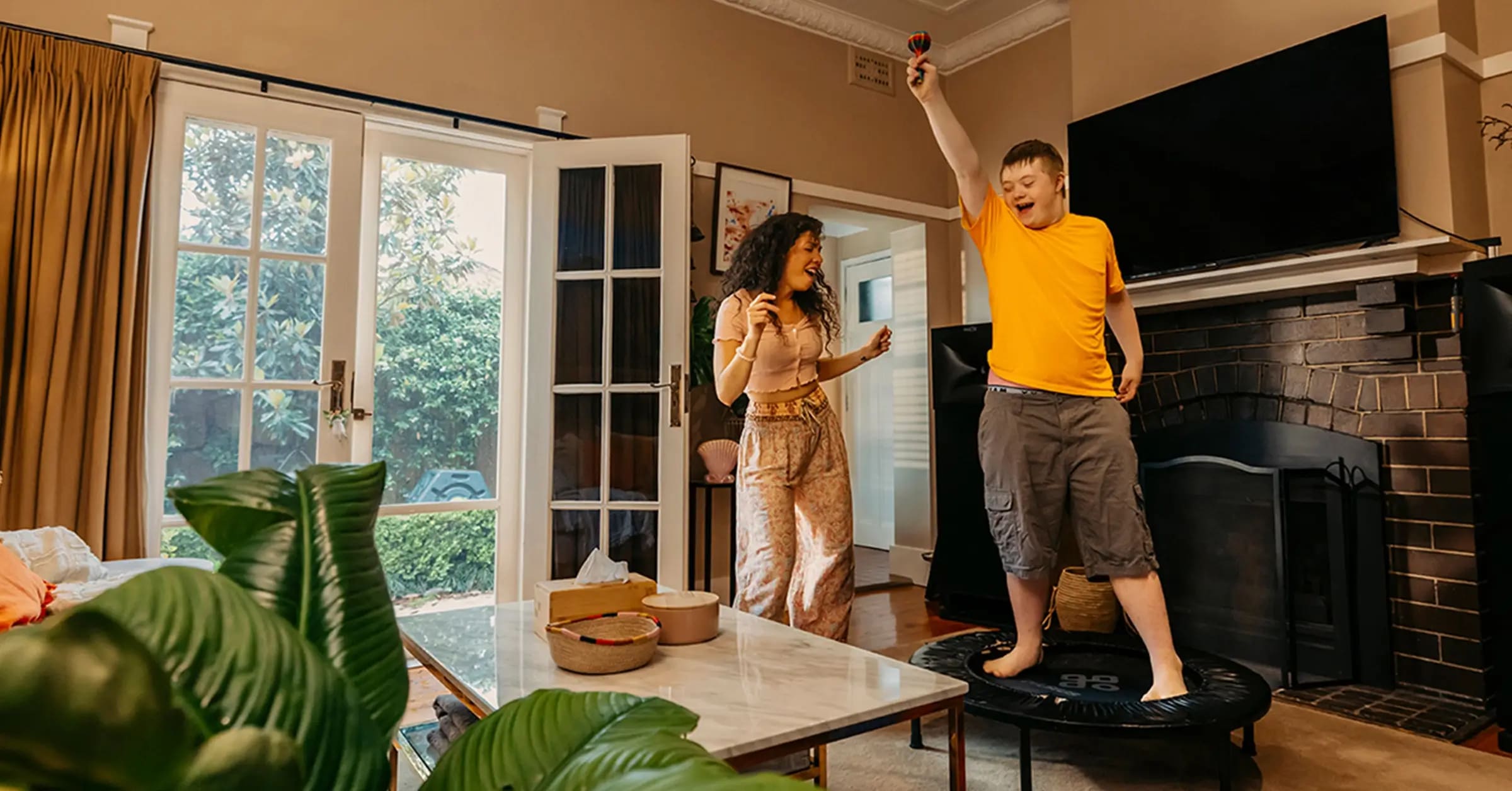 Child with disability stands on mini trampoline and raises a maraca while support worker dances.