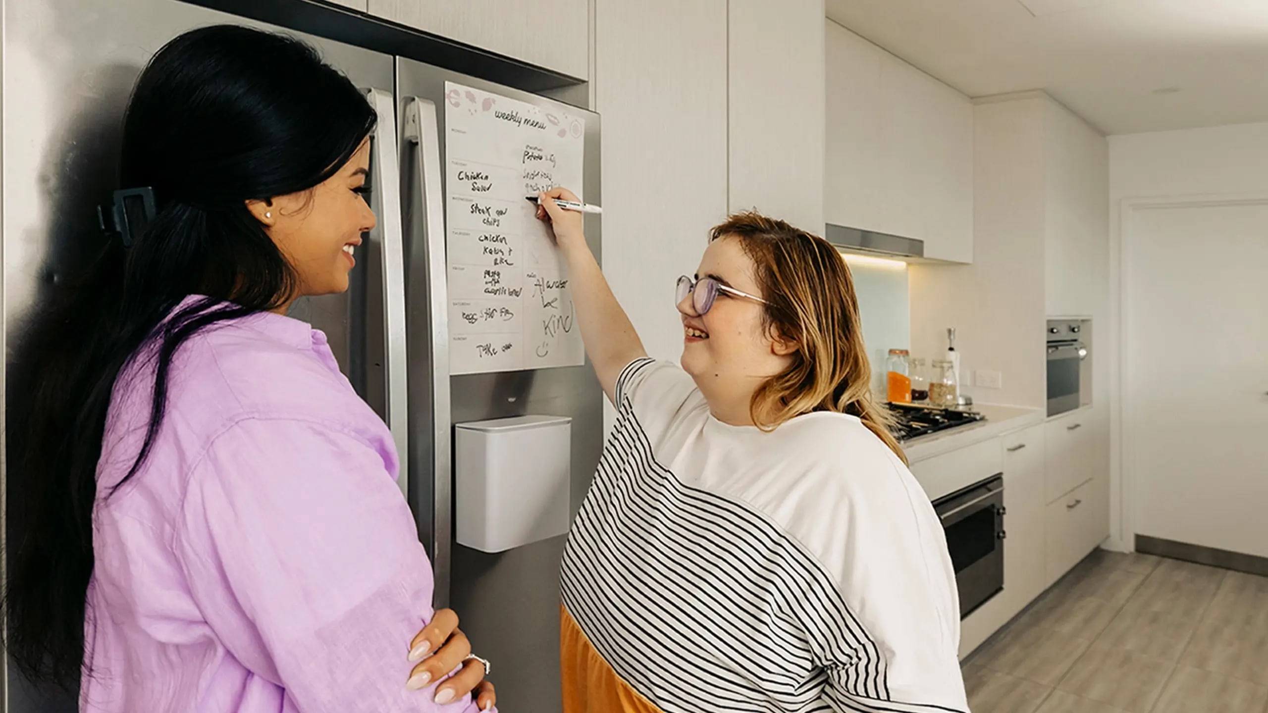 Person with disability in kitchen writes list on fridge while support worker watches.