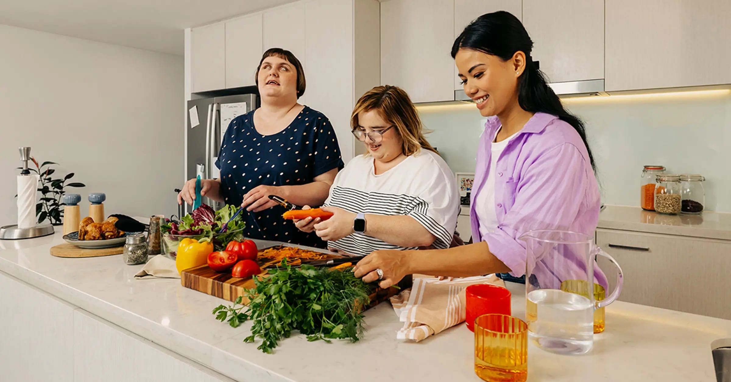 Support worker helps two people with disabilities prepare vegetables at the kitchen bench.