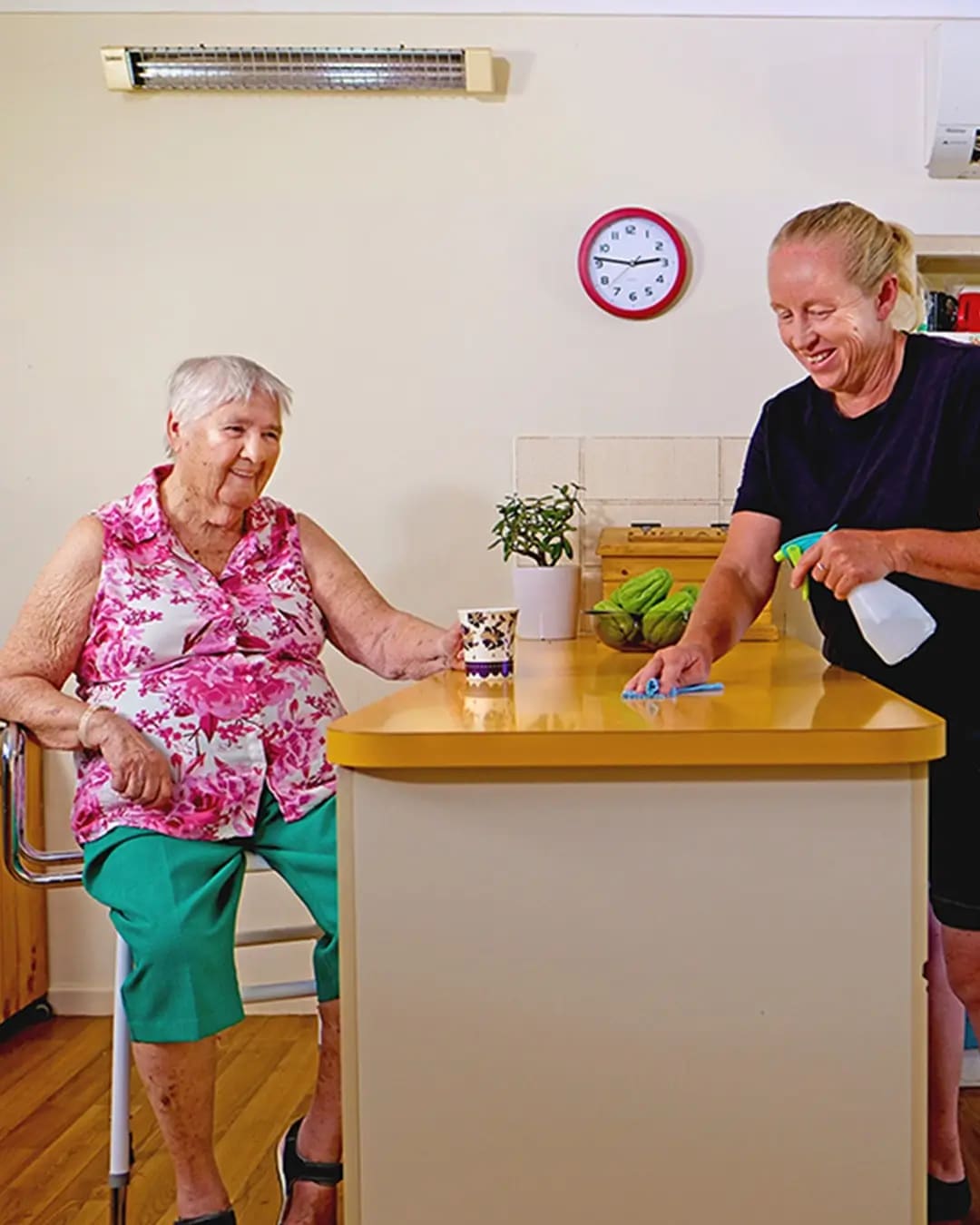 A woman drinks tea while her support worker tidies up.