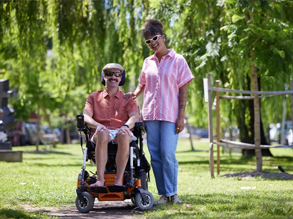 Support worker standing in a park next to client with multiple sclerosis and in wheelchair.