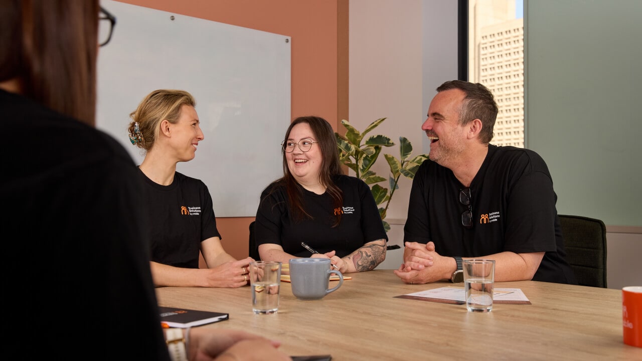 Three smiling colleagues in black 'Business Solutions' t-shirts laughing together around a meeting table.