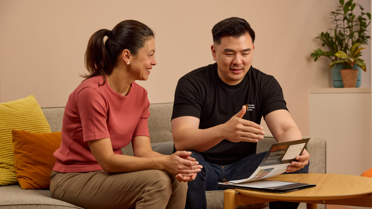 A man in a black t-shirt and a woman in a pink top are seated on a sofa, looking at an open brochure or document on a coffee table, with the man gesturing as he speaks.