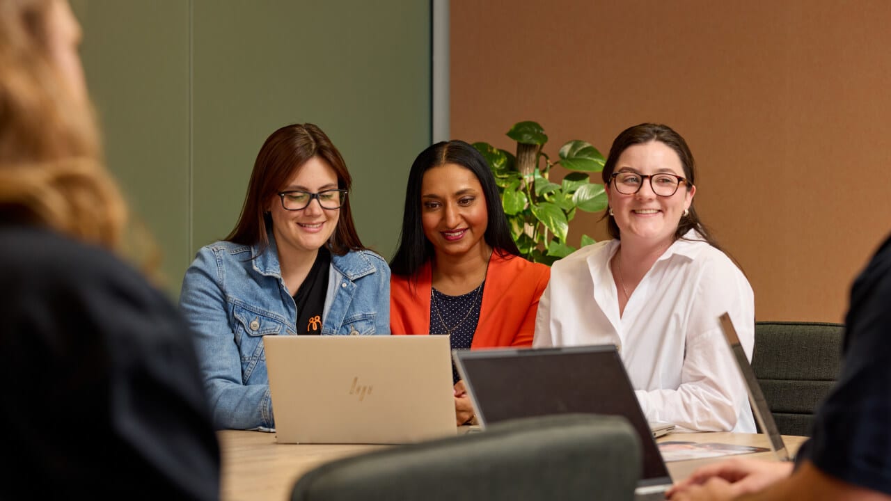 Three smiling women of diverse backgrounds working together on laptops during a business meeting.