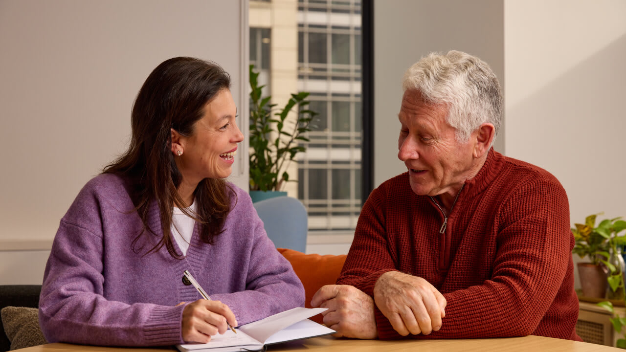 A mature woman in a purple sweater writing in a notebook while talking and smiling with an older man in a red sweater.