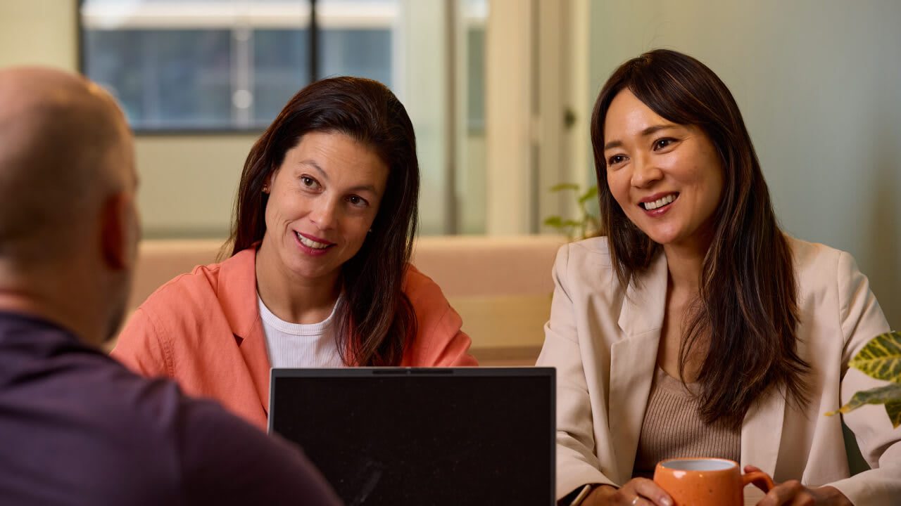 Two smiling professional women, one in an orange blazer and one in a cream blazer, sitting at a meeting table across from an unseen person.