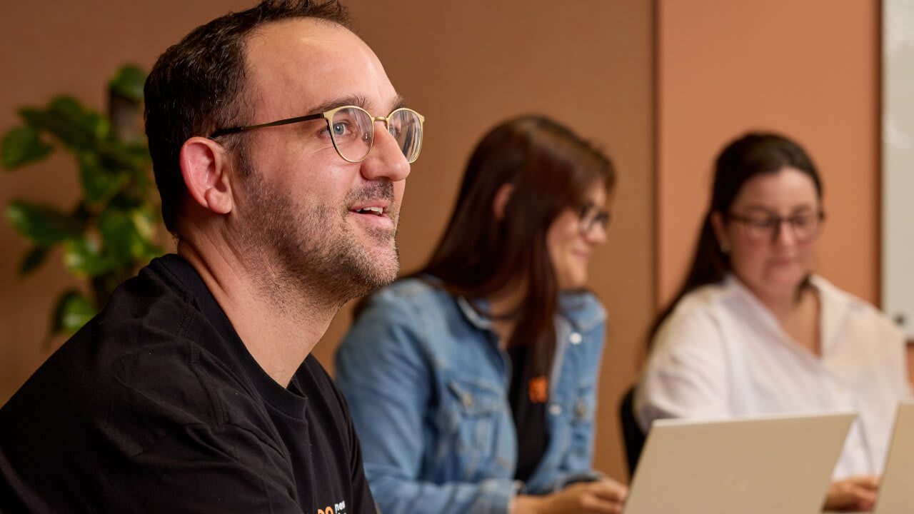 A man in glasses and a black t-shirt smiling while sitting in a meeting with two women in the background.