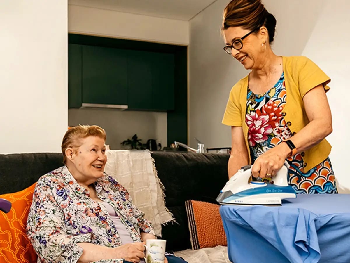 Older person sits on the couch with tea and chats to support worker ironing.