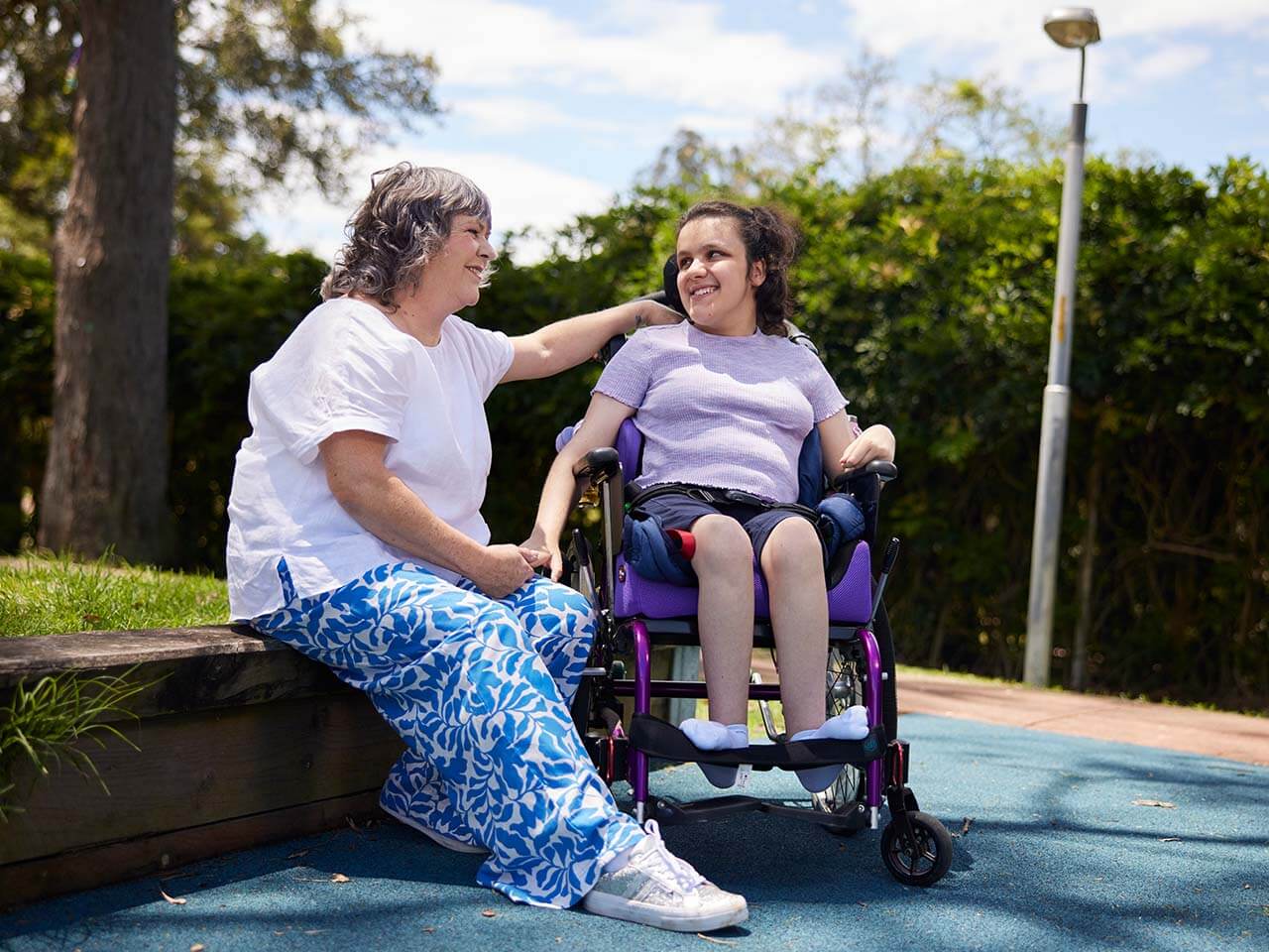 Teenager in a wheelchair in a park with support worker sitting next to them.
