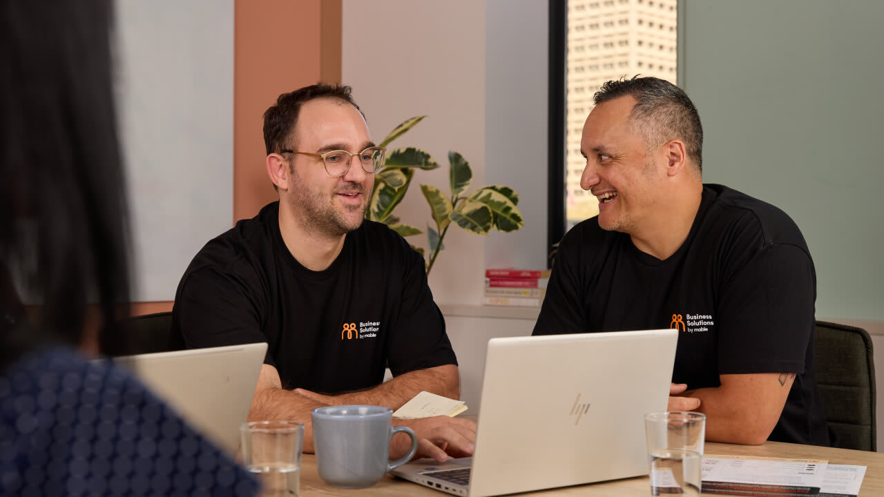 Two smiling male colleagues in black 'Business Solutions' t-shirts sitting across a meeting table with an open laptop, engaged in a discussion.