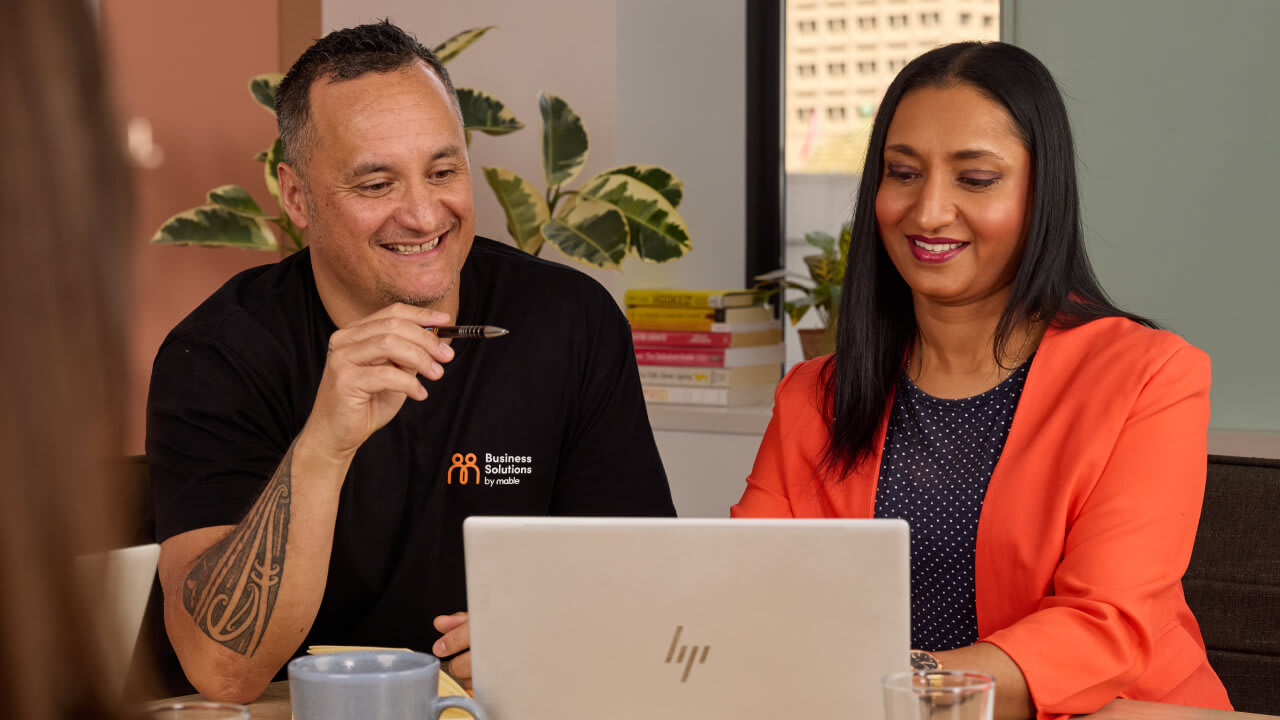 A smiling man in a black 'Business Solutions' t-shirt and a woman in an orange blazer looking down and reviewing content on a laptop screen together.