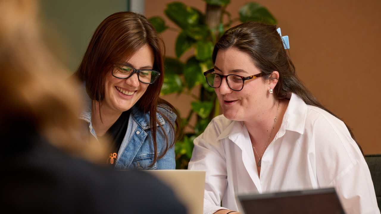 Two smiling women in glasses leaning in together, collaborating and looking at a shared laptop screen during a meeting.