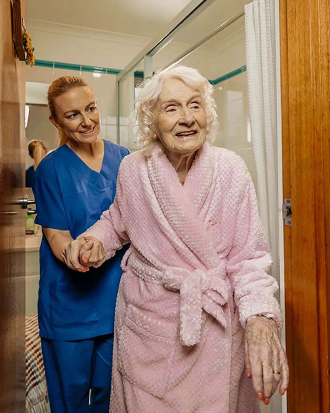 Registered Nurse in scrubs helps older client in pink bathrobe walk out of their bathroom.