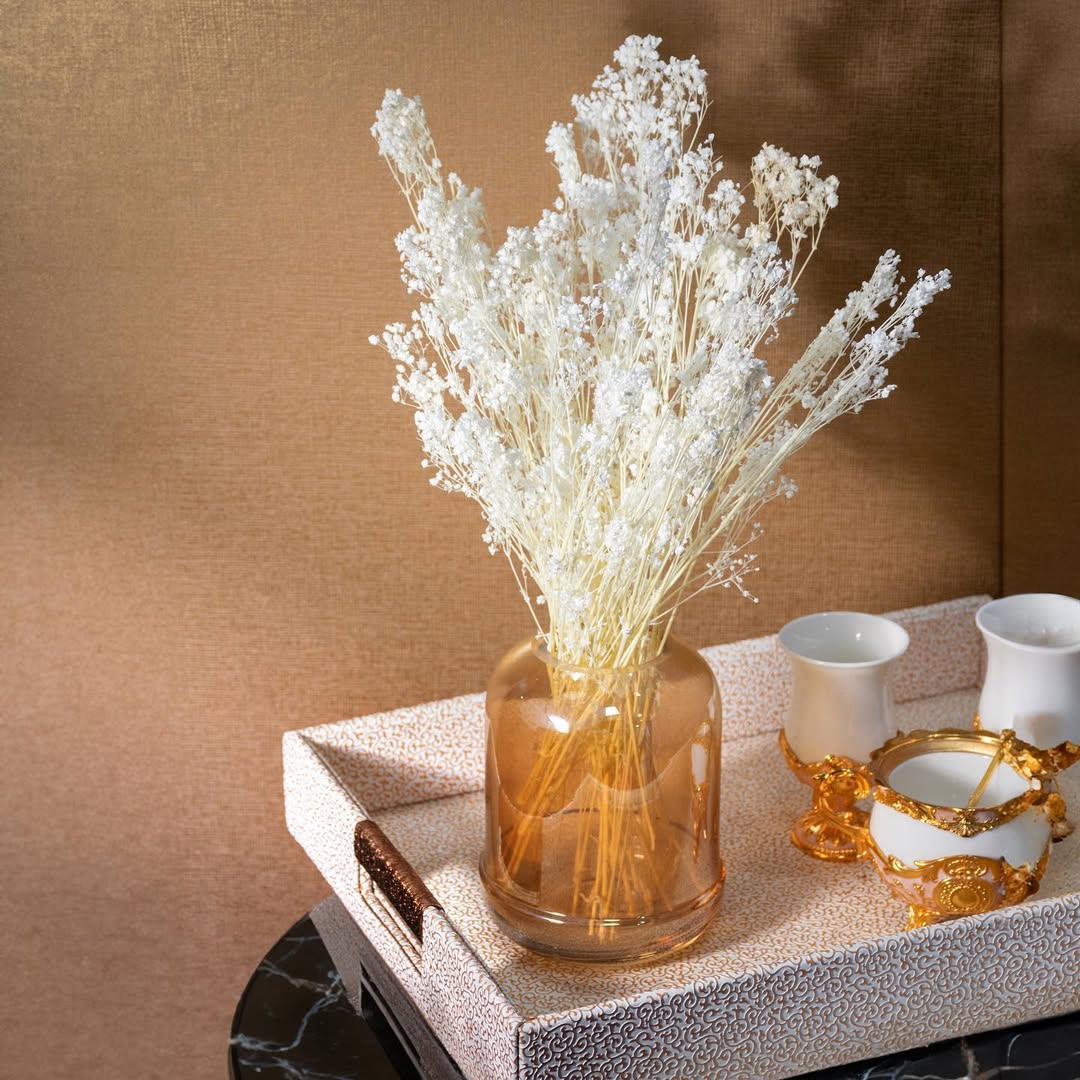 White dried flowers in an amber glass vase with gold-accented white ceramic pieces on a decorative tray