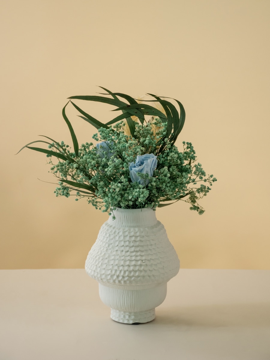 Textured white vase containing a blue rose and green baby's breath with eucalyptus leaves on a beige background