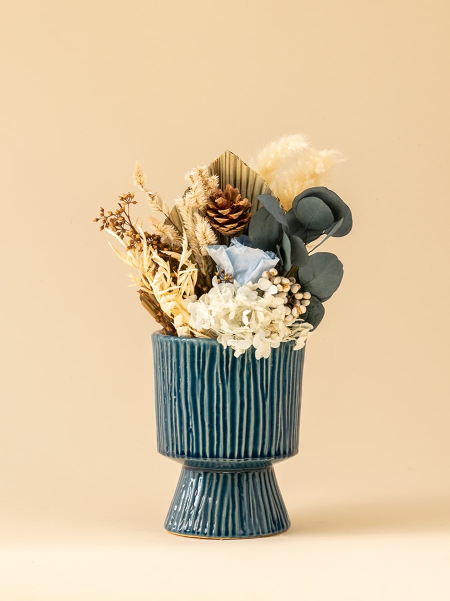 Vibrant dried flower arrangement with white hydrangea, eucalyptus, pine cone and cream bunny tails in a textured blue pedestal vase