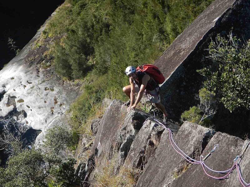 Grimpeur casqué avec un sac rouge progressant sur une arête de roche grise, corde rose, cascade sur une dalle claire en contrebas