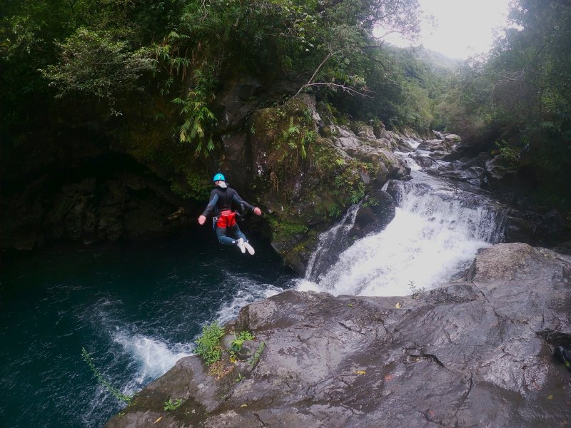 Canyoneur au casque bleu qui saute en faisant un signe de la main au-dessus d'une cascade écumante