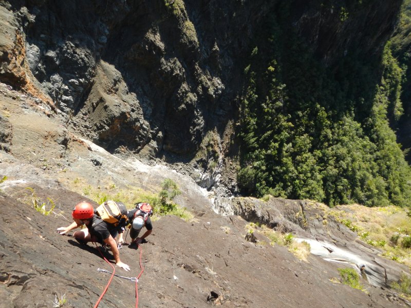 Deux grimpeurs casqués encordés sur une grande dalle de basalte, avec en contrebas une ravine encaissée et une cascade
