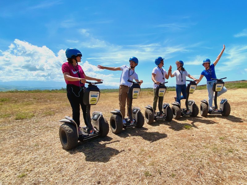Cinq pilotes casqués sur des segways tout-terrain dans une savane rase, deux d'entre eux le bras levé, océan à l'horizon