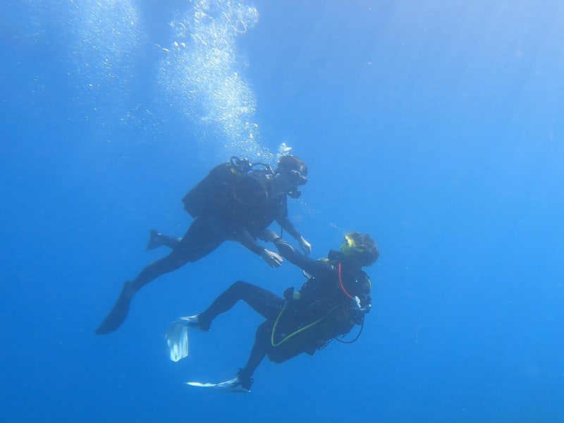 Moniteur qui tient la main d'une élève face à lui, en pleine eau bleue, sous un nuage de bulles