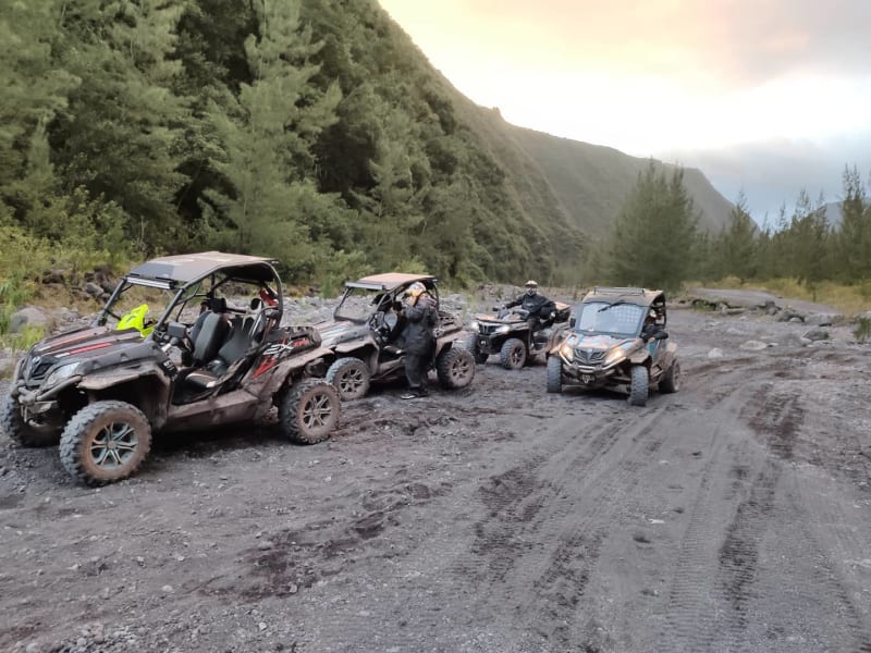 Buggies boueux et un quad arrêtés dans le lit de sable noir et de galets d'une large rivière, pentes boisées autour