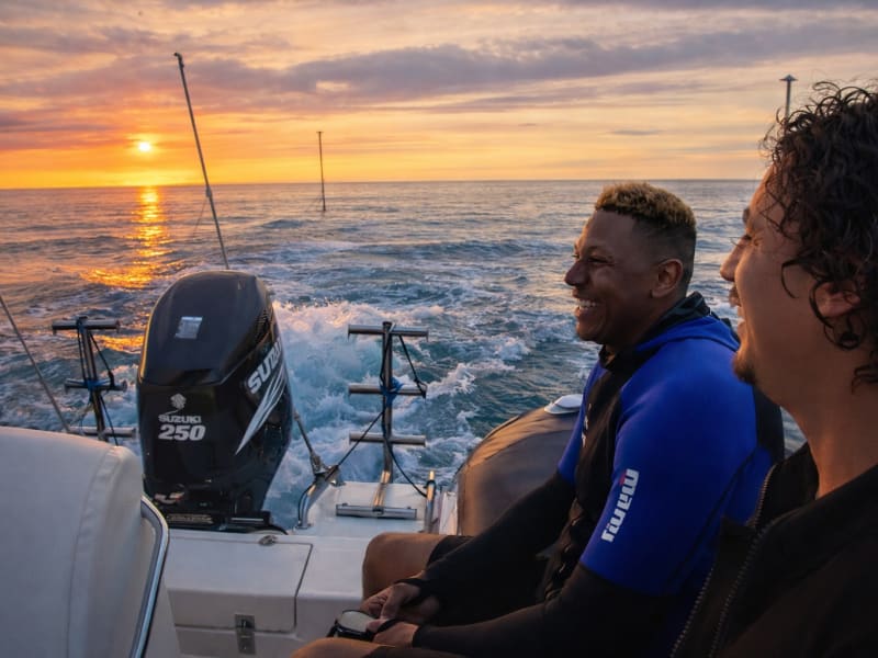 Deux passagers souriants à l'arrière d'un semi-rigide, le soleil couchant sur l'océan derrière eux