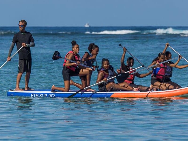 Groupe de jeunes participants sur un paddle géant RTM SUP 18'2 dans le lagon de Trou d'Eau, gilets rouges et moniteur debout, La Saline-les-Bains, La Réunion