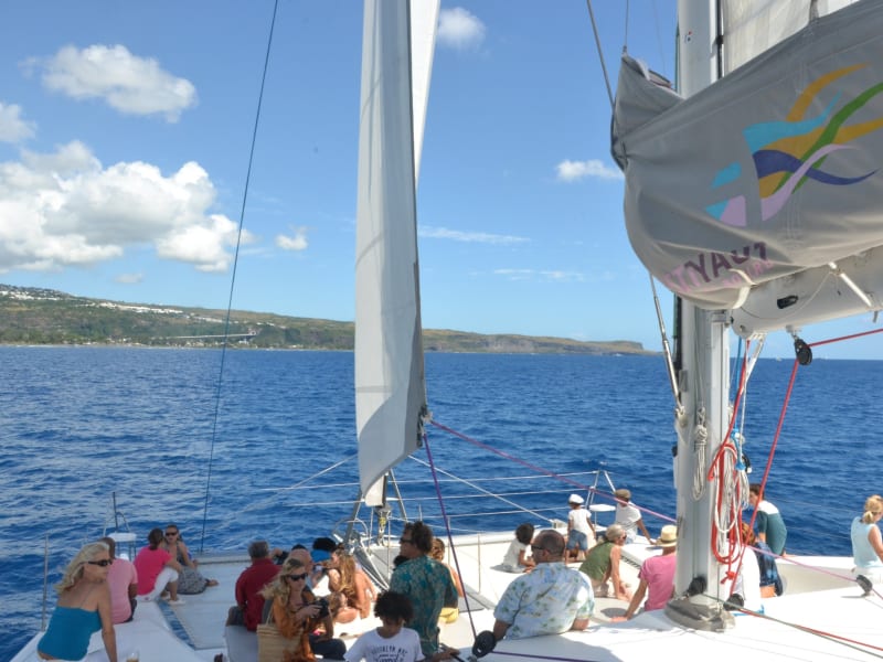 Passagers assis sur le pont avant d'un catamaran sous les voiles, mer bleue et côte verte de l'ouest de La Réunion à l'horizon