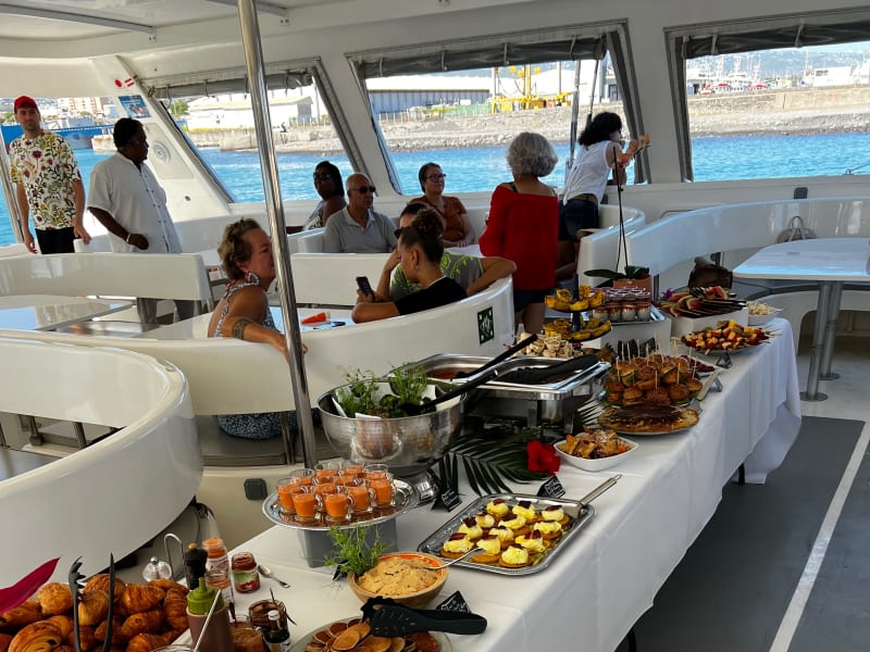 Buffet de brunch dressé dans le salon couvert d'un catamaran, viennoiseries, verrines et plats chauds, passagers attablés et port en arrière-plan