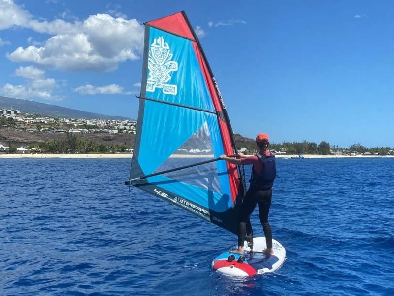 Windsurfeur en cours de planche à voile dans la baie de Saint-Gilles, voile Starboard rouge et bleue, plage et littoral de l'ouest de La Réunion en arrière-plan