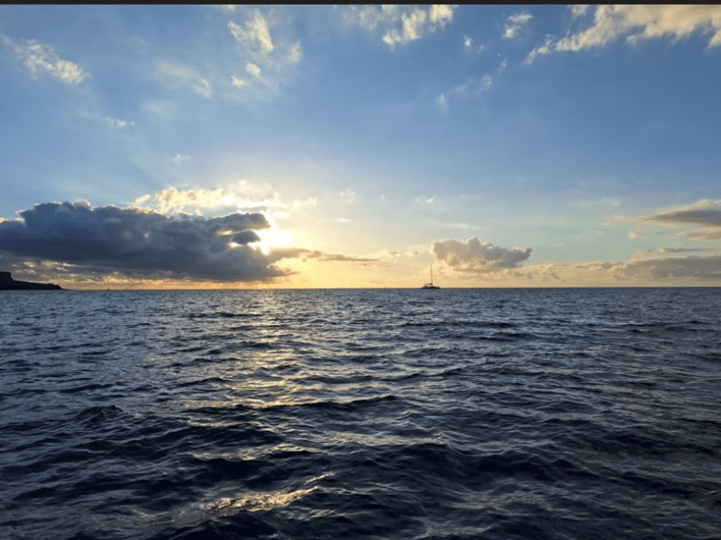 Soleil déclinant derrière un banc de nuages au-dessus de l'océan, un voilier au loin sur l'horizon, vu depuis la mer au large du Port