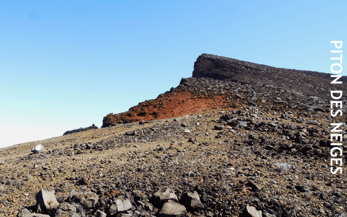Sentier rocailleux conduisant au sommet du Piton des Neiges, croupe minérale rouge et grise du cône terminal, ciel bleu, La Réunion