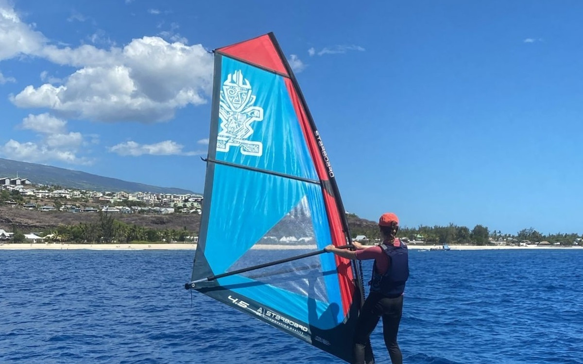 Windsurfeur en cours de planche à voile dans la baie de Saint-Gilles, voile Starboard rouge et bleue, plage et littoral de l'ouest de La Réunion en arrière-plan