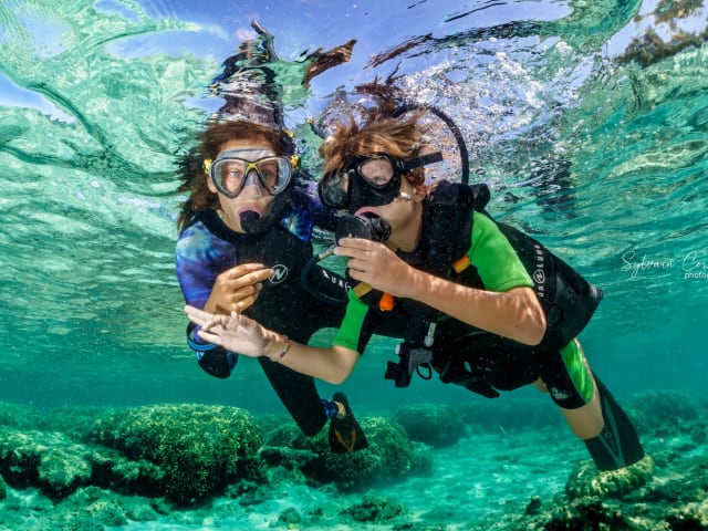 Deux enfants équipés de masques et de détendeurs, sous la surface d'une eau turquoise très claire, l'un fait le signe OK