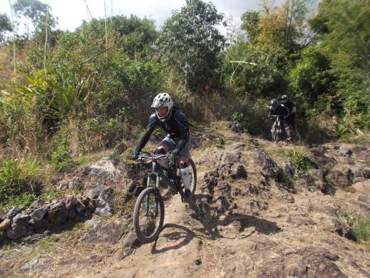Pilote en casque intégral qui franchit un passage de roche et de terre sur un VTT de descente, d'autres pilotes derrière