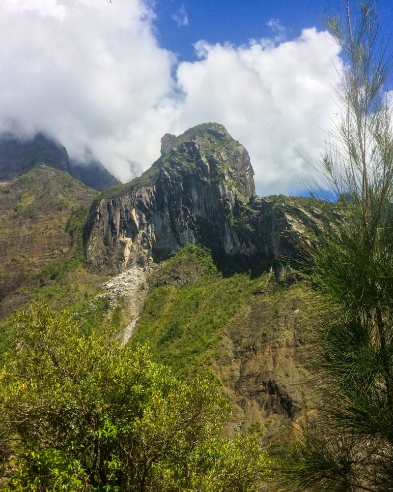 Piton rocheux aux flancs gris et coulée d'éboulis clair, crêtes dans les nuages, filao au premier plan