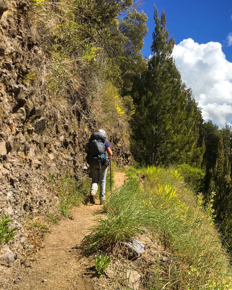 Randonneur sur un sentier étroit à flanc de rempart, face à un piton pointu couvert de forêt