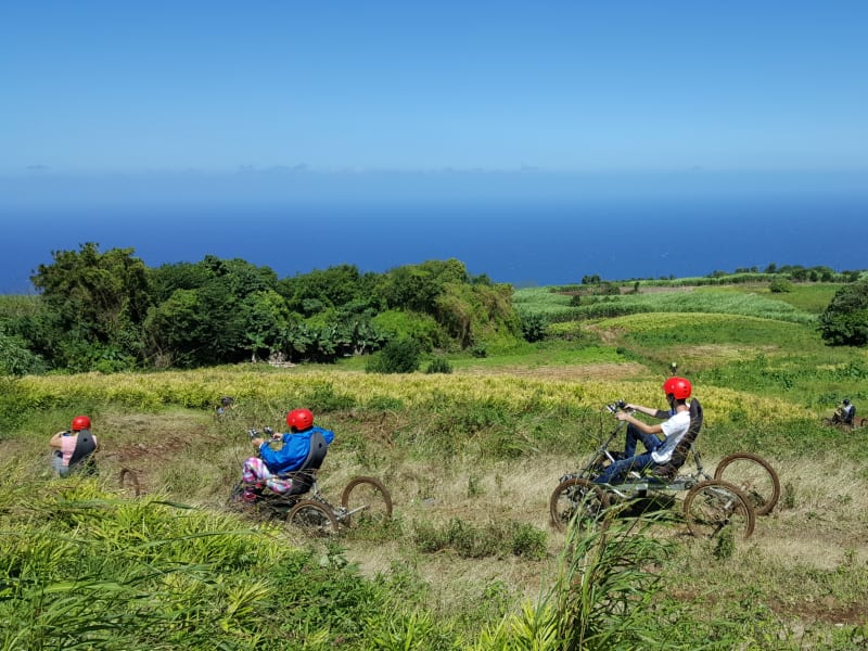 Trois quadbikes pilotés par des personnes casquées de rouge descendant une prairie en pente, océan et ciel bleu à l'horizon