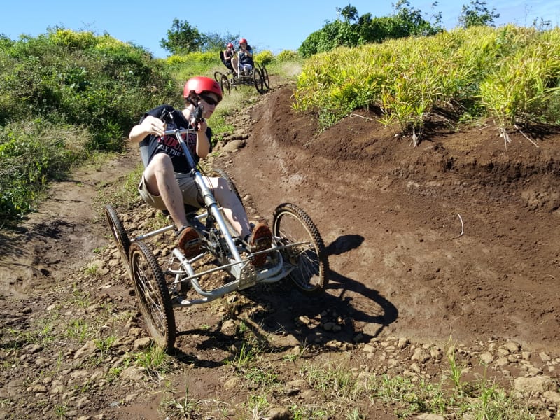 Pilote casqué de rouge sur un quadbike dans une ornière de terre, un second pilote plus loin sur la piste, végétation autour
