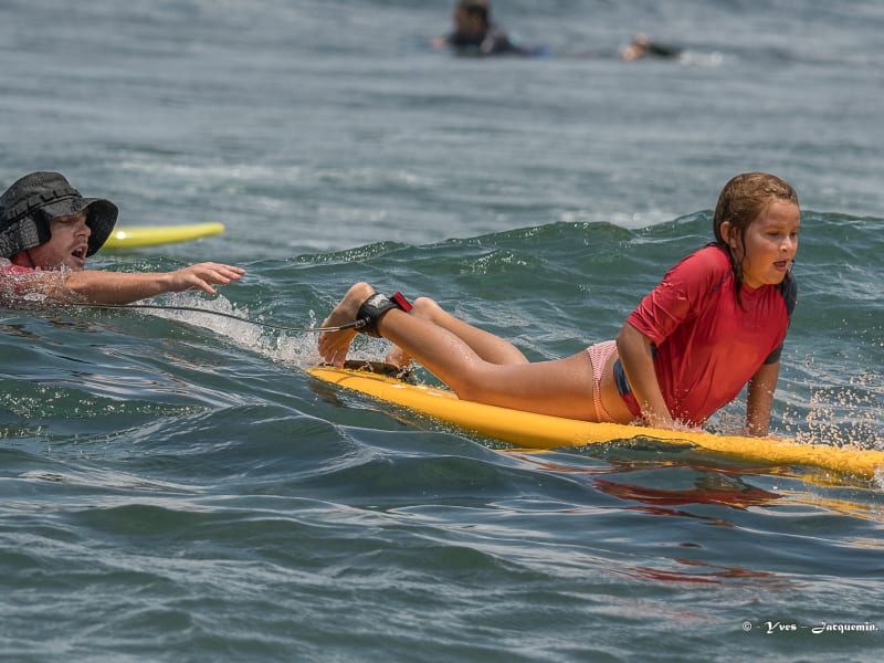 Petite fille en lycra rouge allongée sur une planche jaune qui part sur une vague, moniteur casquetté dans l'eau qui vient de la pousser
