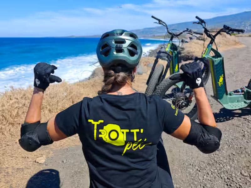 Pilote vue de dos, bras levés, tee-shirt TROTT'PÉI, casque et coudières, trottinettes tout-terrain garées sur un chemin côtier au-dessus de l'océan