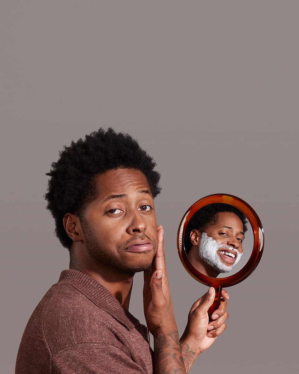 Black male model holding a mirror with an image of his face using MANSCAPED® Beard Conditioner