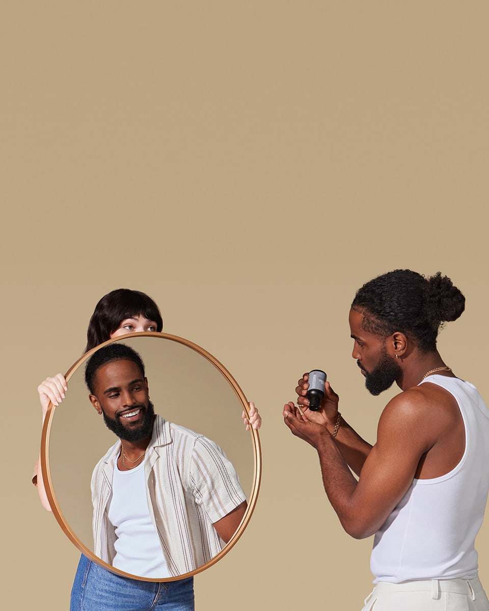 Female model holding up mirror for black male model to help apply Beard Shampoo