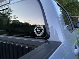A white sticker on the Tacoma’s rear window that has a six-gear shift pattern and the words The Manual Gearbox Preservation Society circling it.