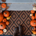 Bird's eye view of brown welcome mat surrounded by mini pumpkins with brown boots walking over it