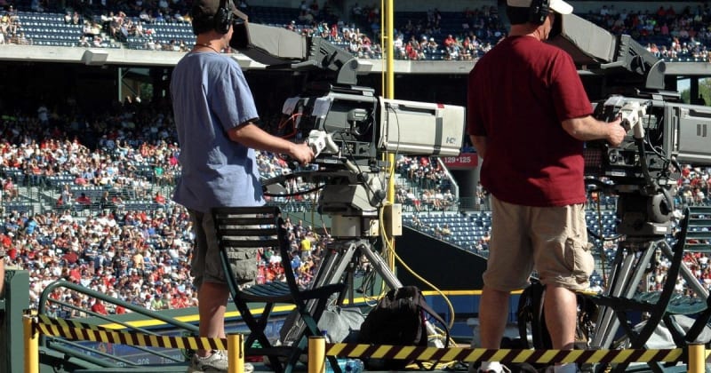 Camera operators capturing a live sports event at a crowded outdoor stadium filled with spectators.