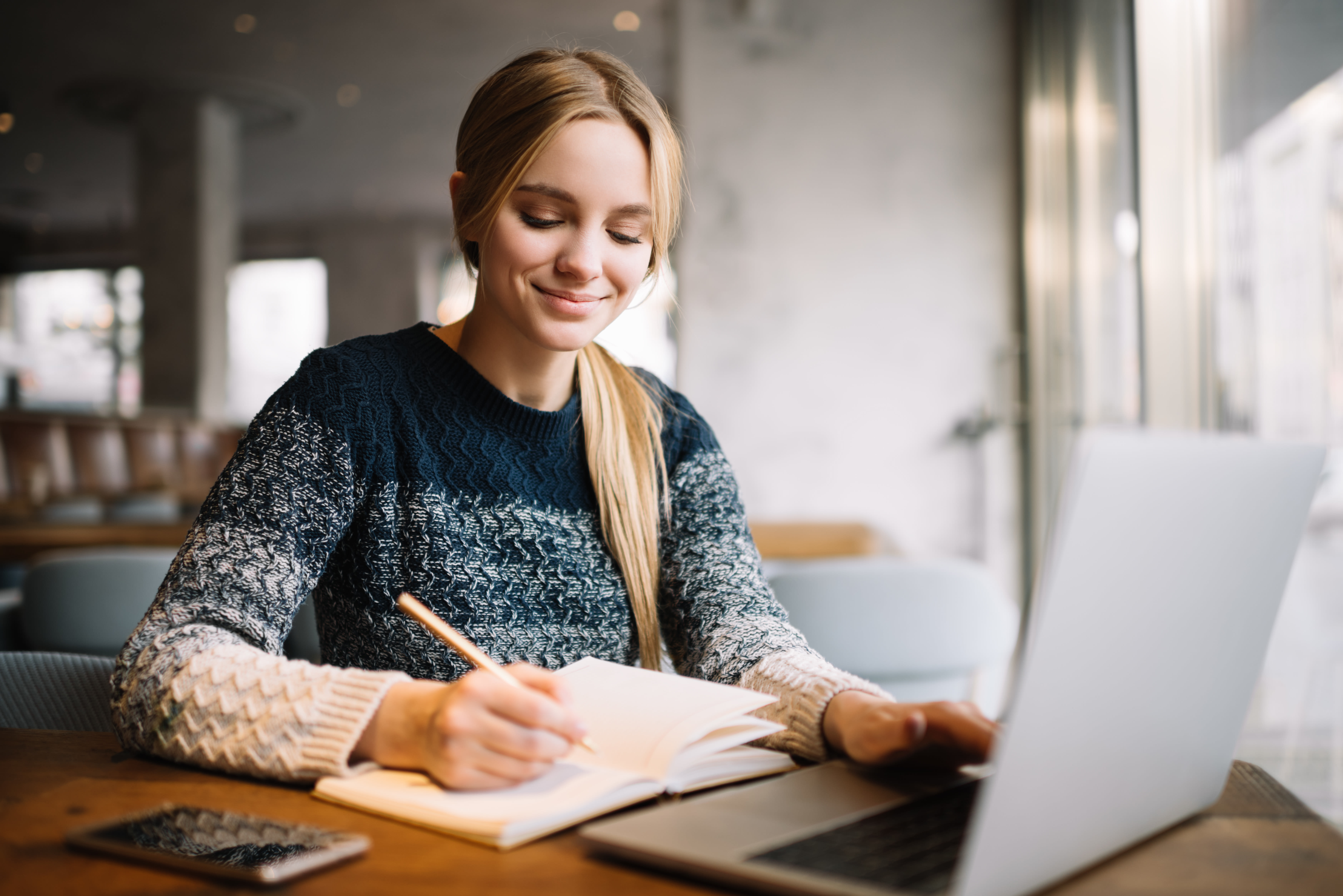 Young attractive university student using laptop computer, studying at modern library. Cheerful caucasian woman writes notes, planning working process, sitting in loft cafe. Exam preparation.