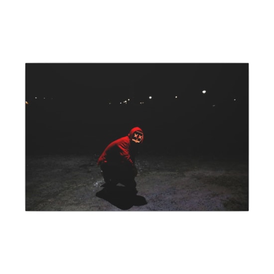A person in a red hoodie with a glowing mask crouches in the shadows on a dark night. Eerie setting.