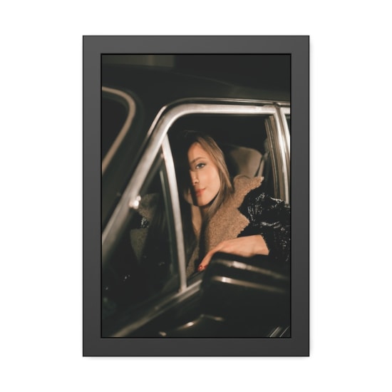 A fashionable woman sits in a vintage car, partially illuminated under soft lighting.